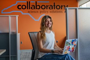 Rose Houck poses for a portrait in front of an orange wall at the NC Collaboratory office in Kenan Labs.